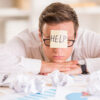 A frustrated man with a "HELP" sticky note on his forehead sits at a messy desk with crumpled papers and charts, looking overwhelmed with bookkeeping tasks