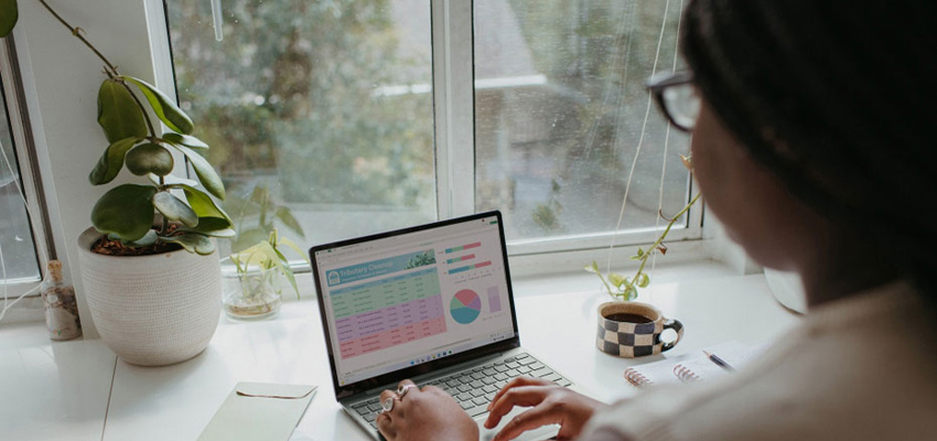 A bookkeeping assistant working on a laptop preparing reports, sitting by a window with a plant and a coffee mug nearby