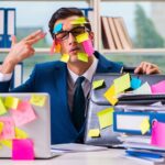 A stressed executive in a suit sits at a cluttered desk covered in sticky notes, files, and papers, looking overwhelmed with tasks.
