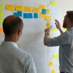 Two men in an office brainstorming, placing colorful sticky notes on a whiteboard planning virtual assistant onboarding