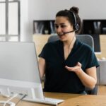 A virtual assistant wearing a headset sits at a desk, speaking and gesturing during an online meeting in a home office