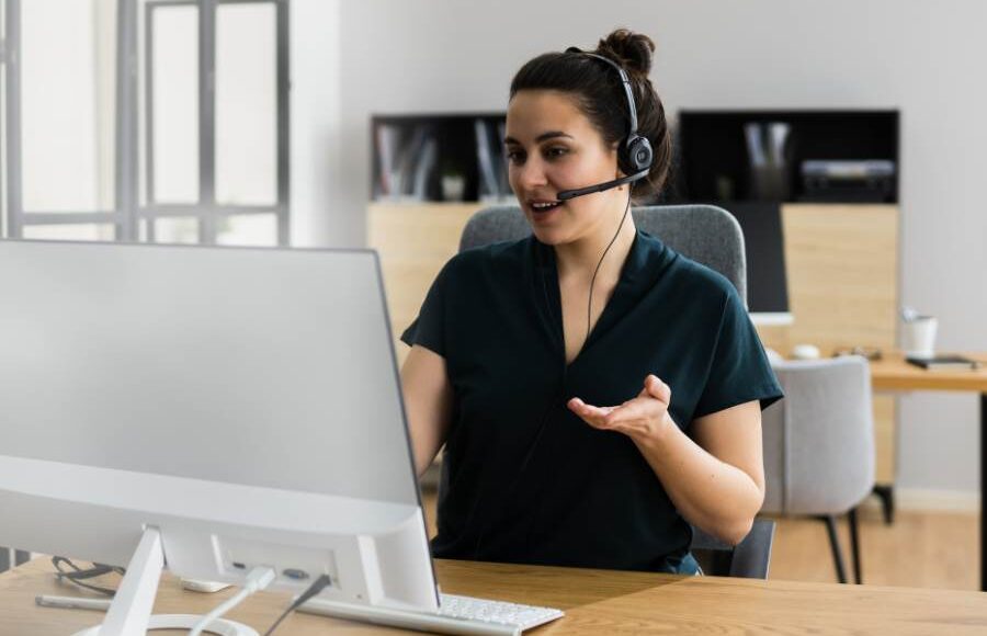 A virtual assistant wearing a headset sits at a desk, speaking and gesturing during an online meeting in a home office